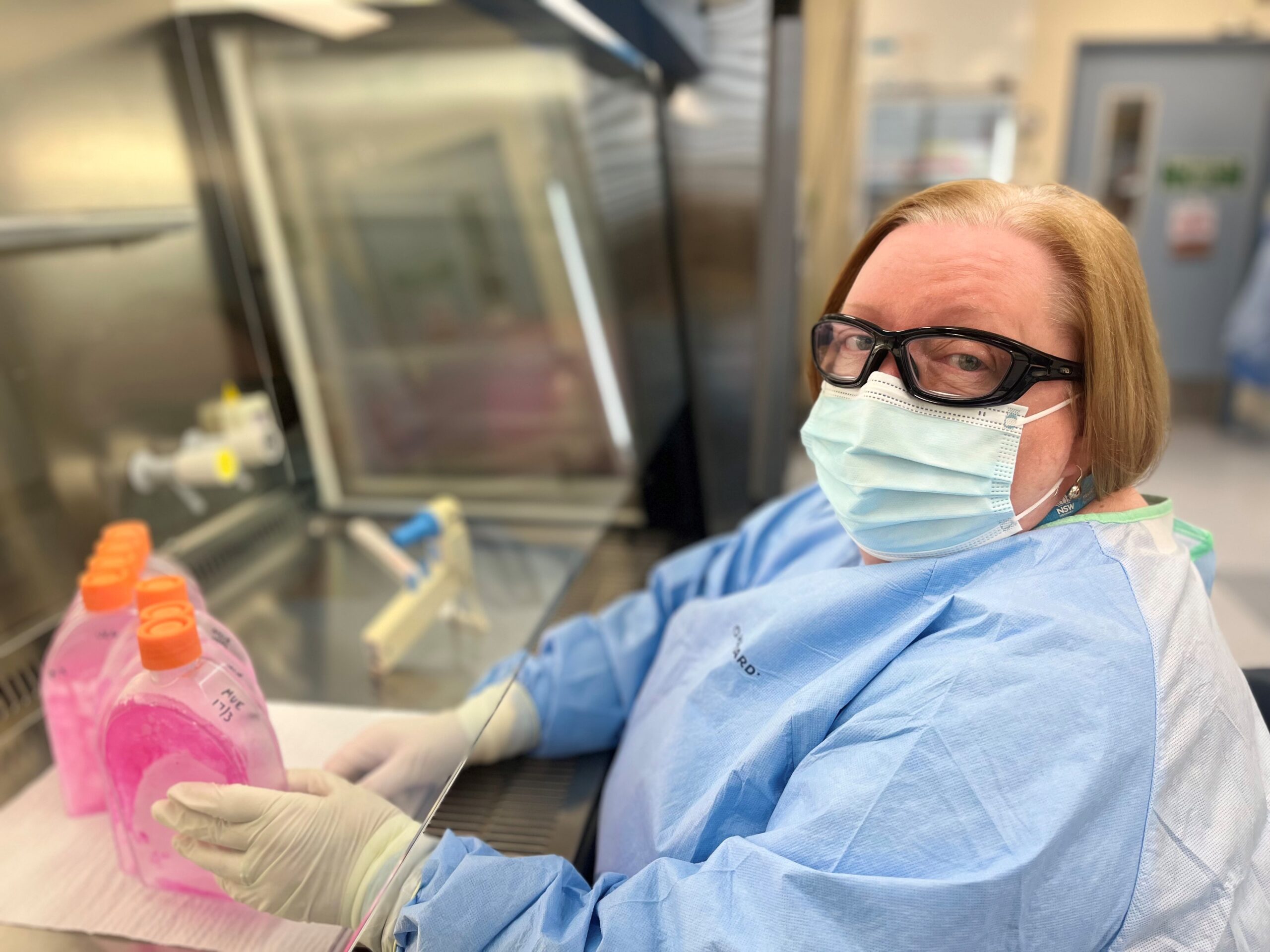 A woman wearing a mask, protective glasses and a protective gown sits in a lab holding containers of pink liquid.