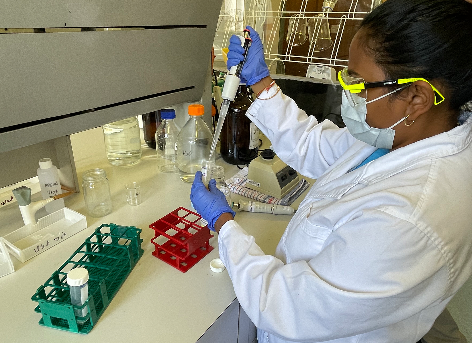 A woman in a lab coat and safety glasses using a pipette and test tubes.