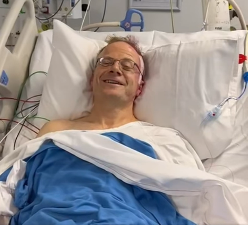 A man lying in a hospital bed, with the side of his head bandaged, smiling.