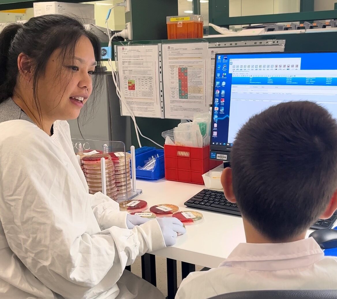 A woman wearing a white lab coat sits at a laboratory desk talking to a young boy in a lab coat.