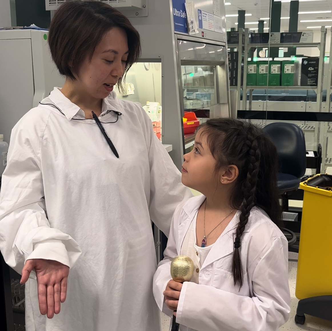 A woman in a white lab coat stands next to a young girl in a lab coat holding a microphone.