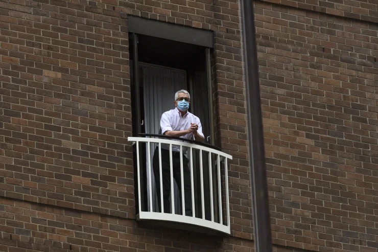 A man wearing a surgical face mask leans on a balcony railing in a high-rise brick building.