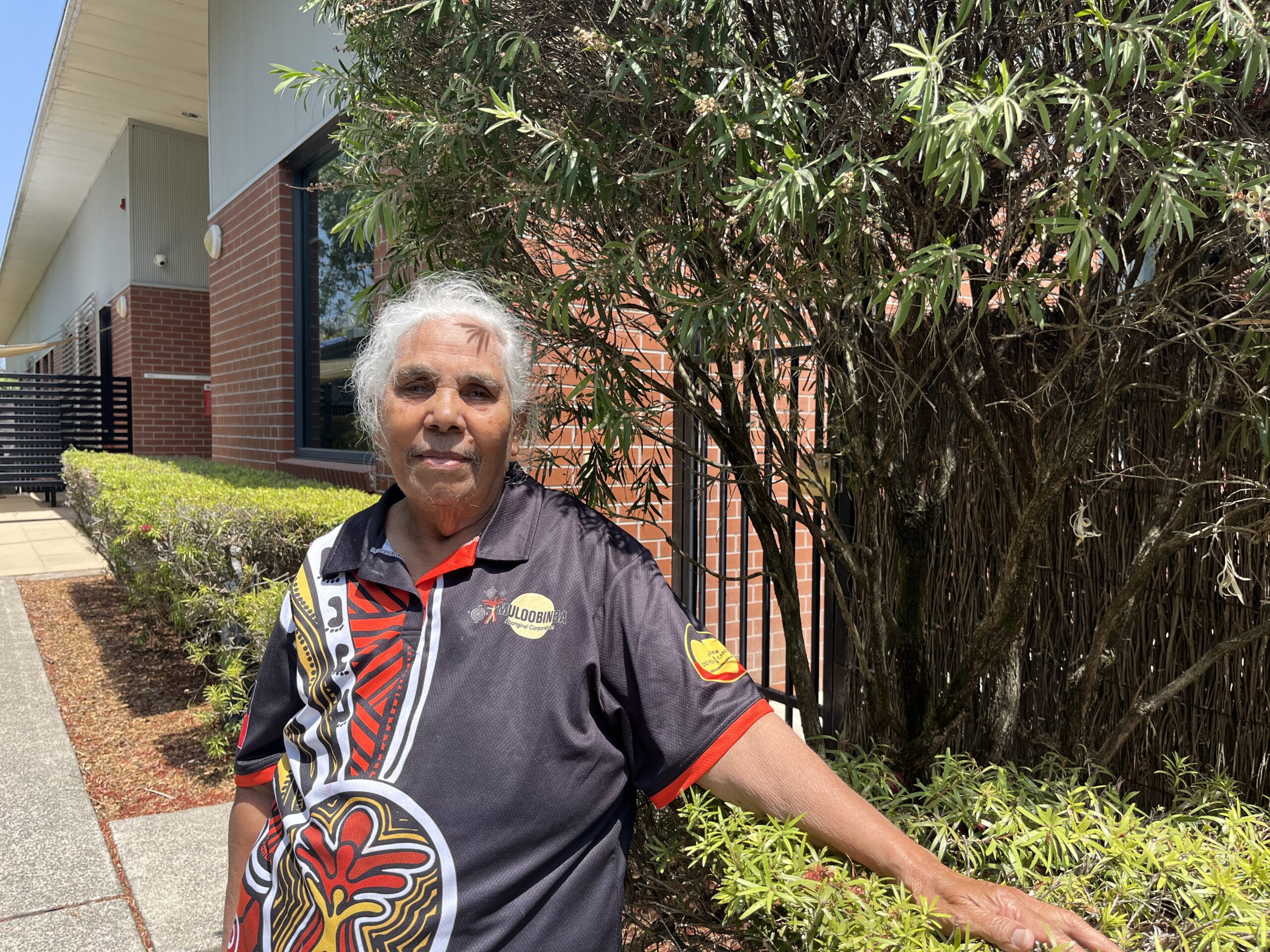 An elderly First Nations woman stands in a garden wearing a black shirt with red, yellow and white Aboriginal designs.