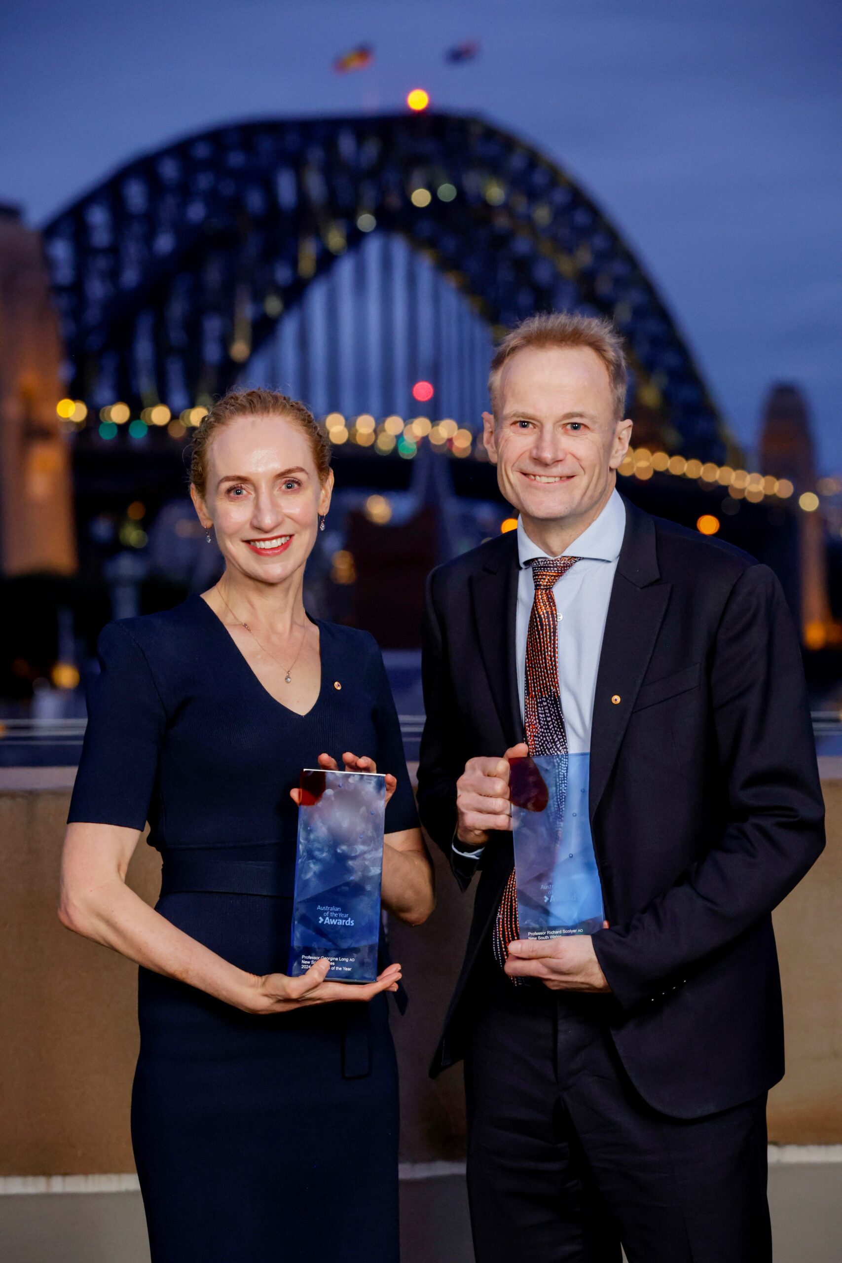 A man and a woman holding trophies stand in front of the Sydney Harbour bridge at night.