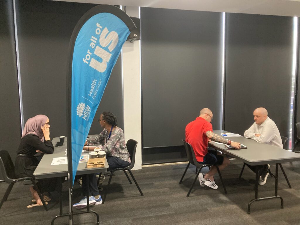 People sitting at tables talking, near a NSW Health Pathology banner.