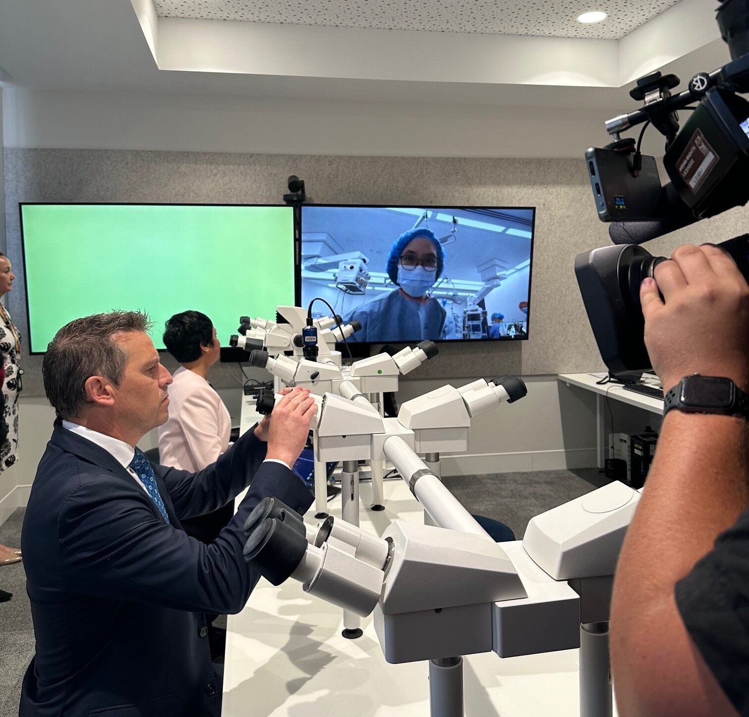 The Minister looking at a microscope, while a surgeon appears on a video screen in the background.