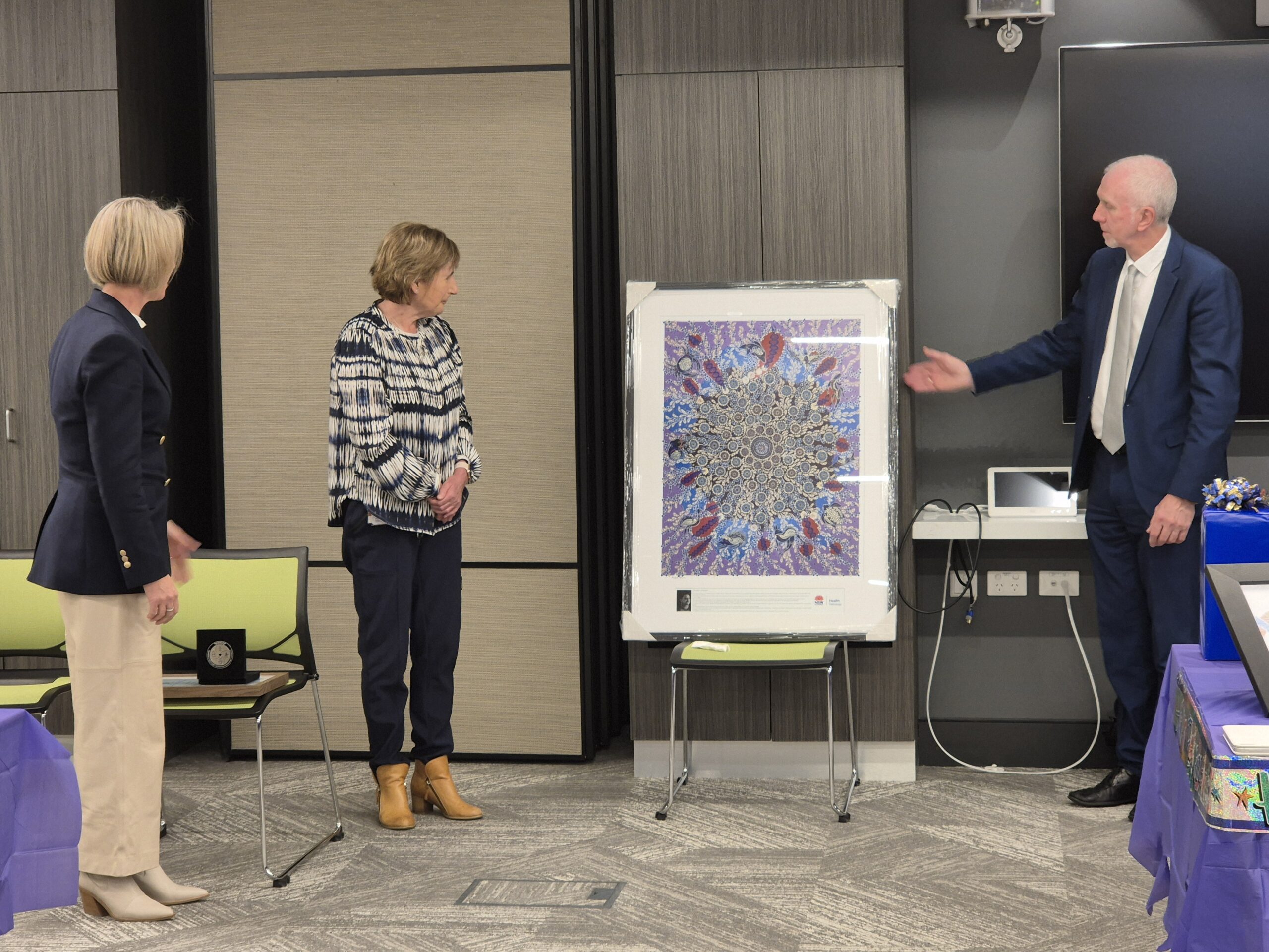 Two women and a man standing in a large room looking at an Aboriginal artwork.