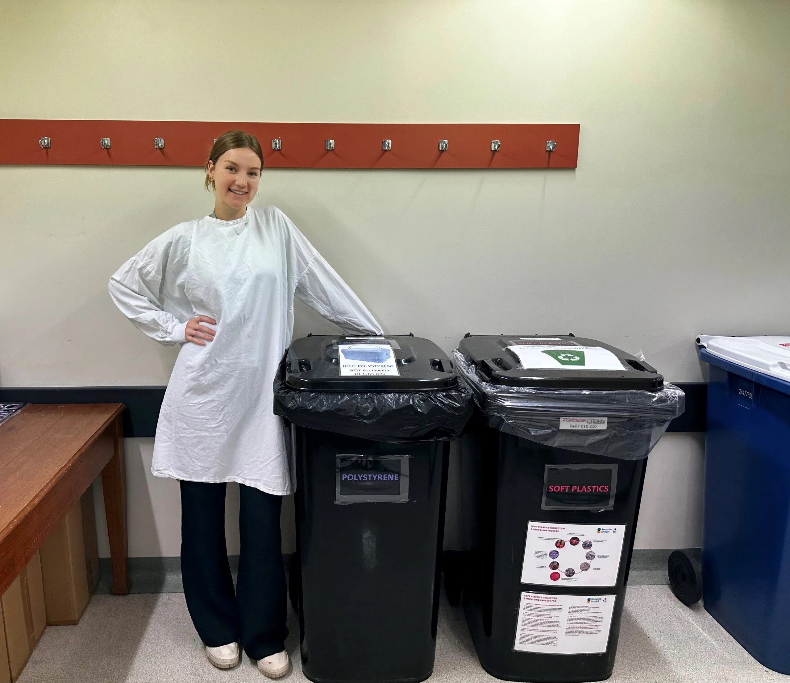 A woman in a white lab coat, leans on a large black recycling bin.