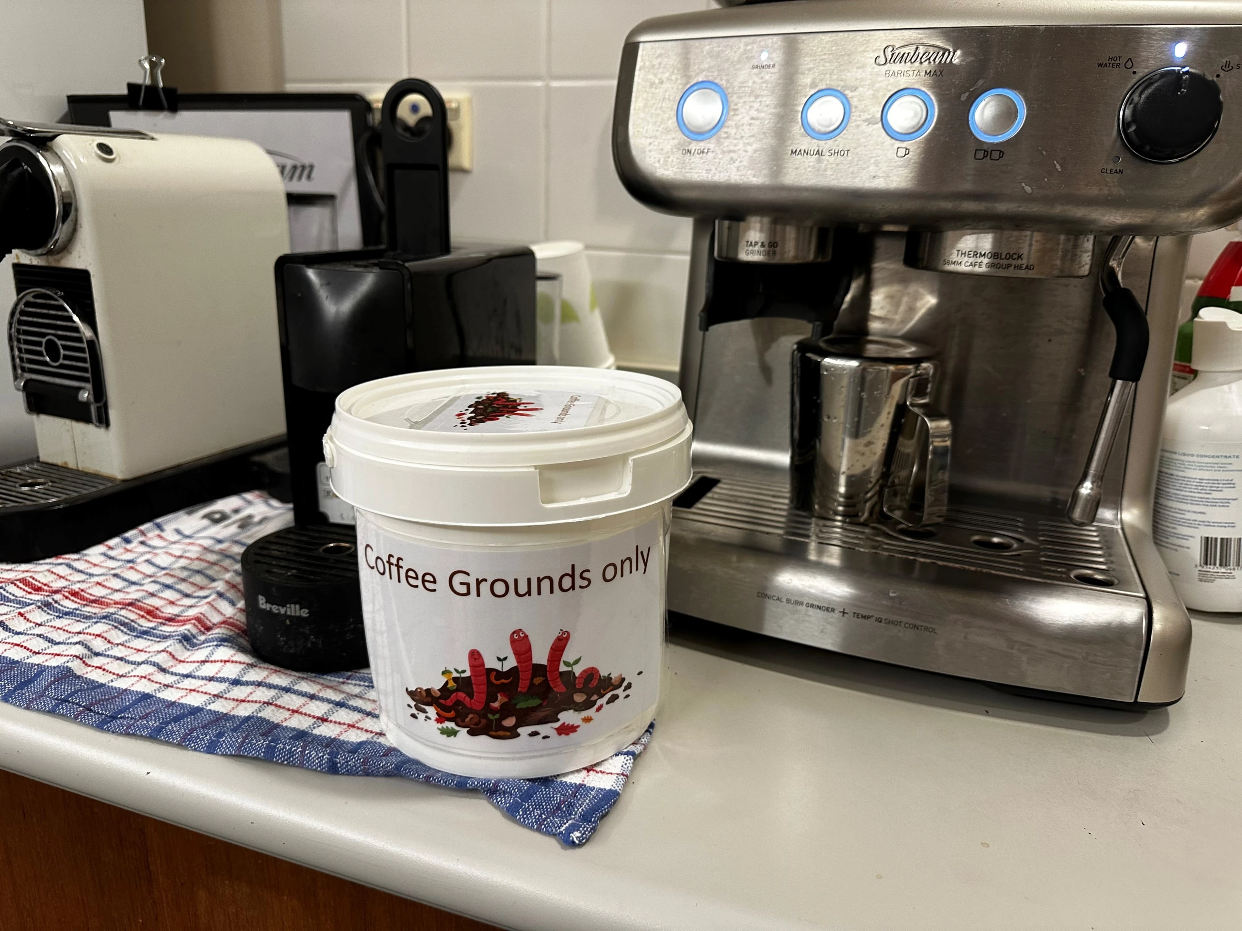 A bucket labelled "Coffee Grounds Only" on a kitchen bench near a coffee machine.