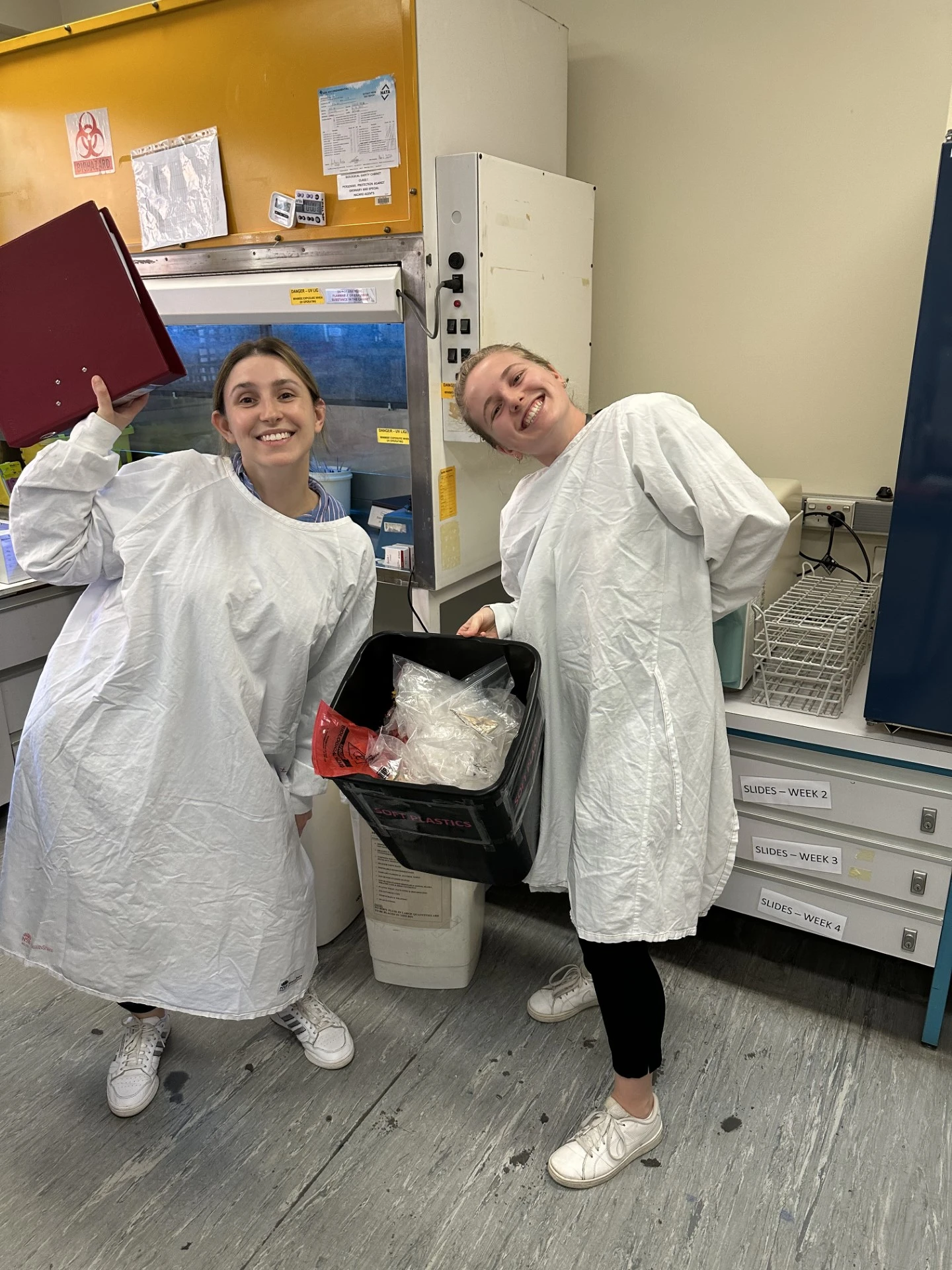 Two young women wearing white lab coats holding up a soft plastic recycling bin.