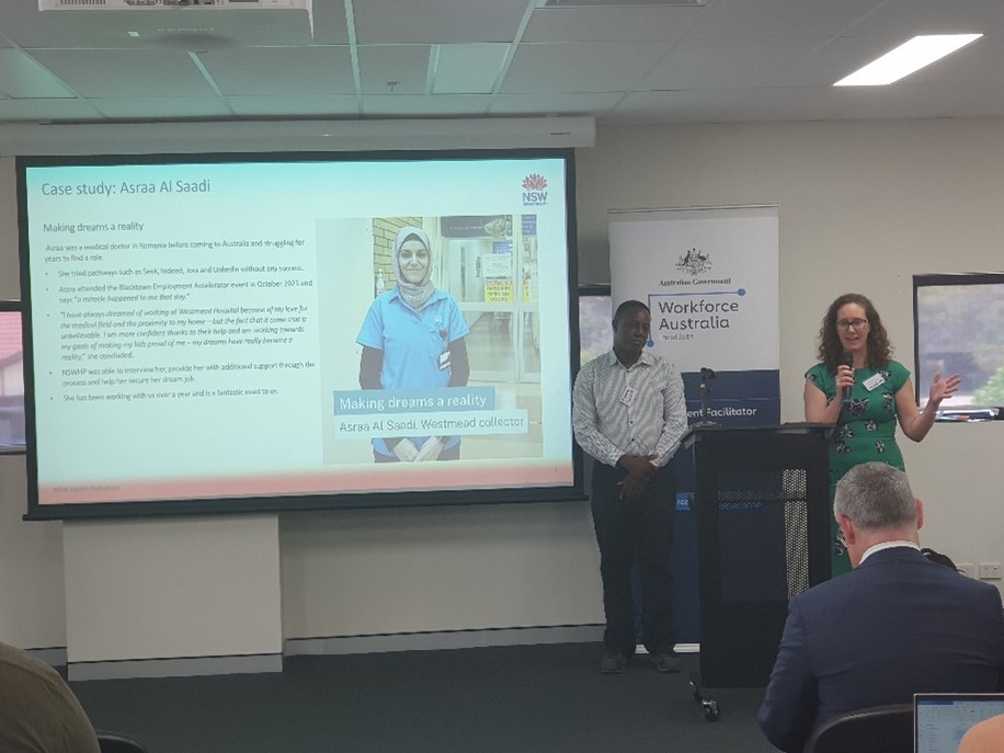 A woman stands at a podium giving a speech, with a photo of a health worker on a screen in the background.