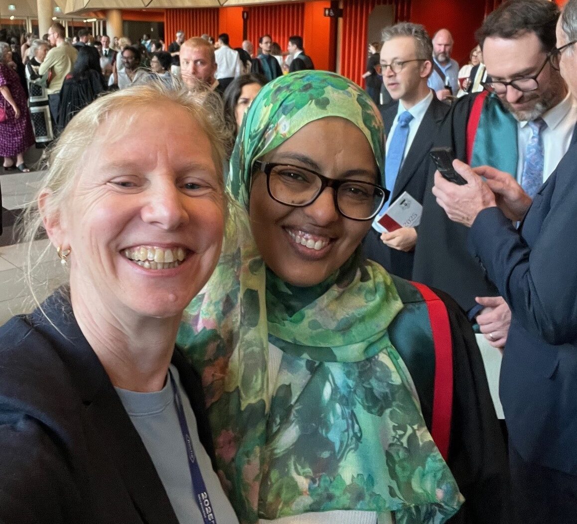 Two women, one in graduation robes, smiling in a roomful of people.