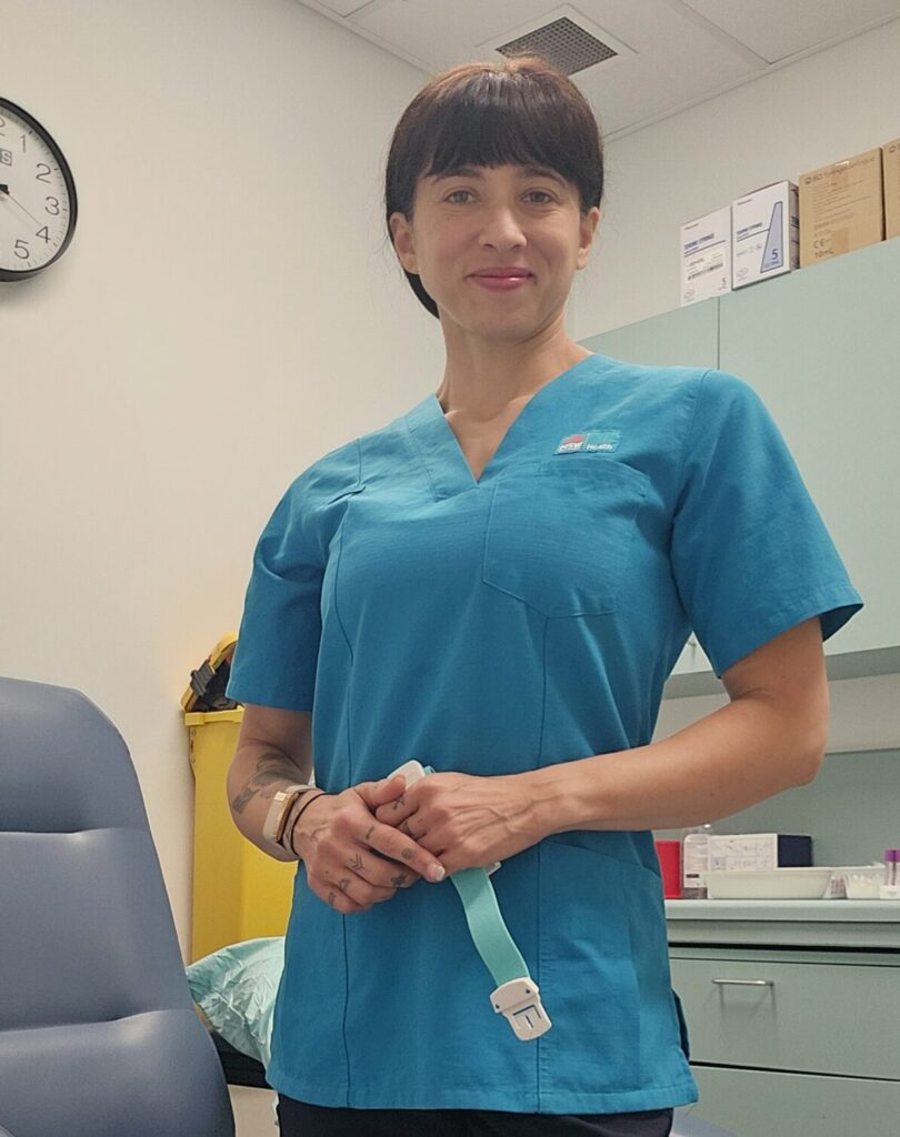 A woman wearing a healthworker uniform, smiling while standing in a collections space.