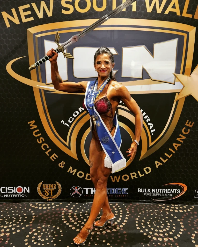 A female bodybuilder wearing a blue sash and holding a sword aloft.