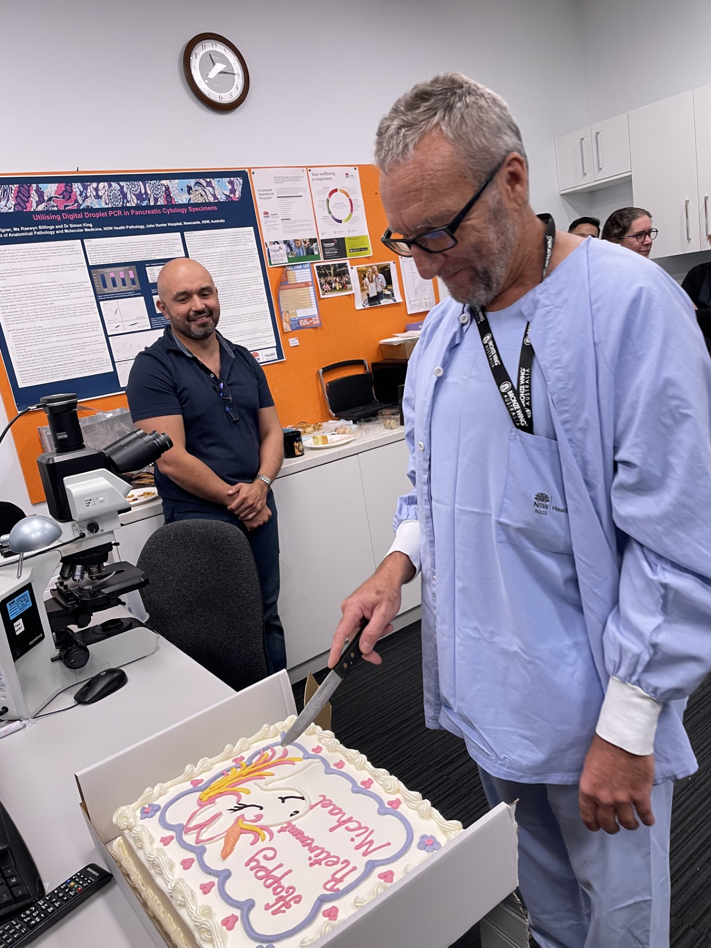 A man in blue hospital scrubs cuts a large cake.
